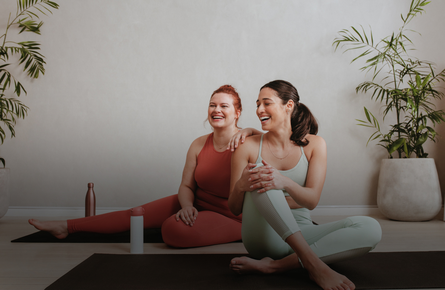 Zwei Frauen sitzen auf Yogamatten in Sportklamotten und lachen